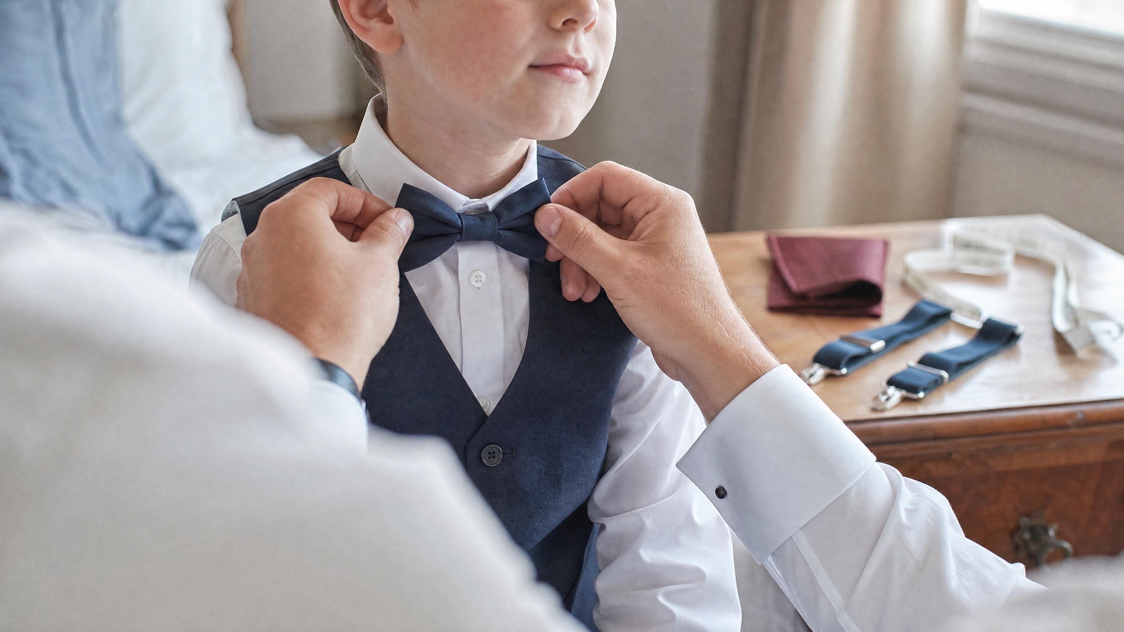 Un adulte ajuste un nœud papillon sur un enfant en chemise blanche, avec un mètre ruban visible sur une commode, dans une chambre lumineuse avant une cérémonie Billede